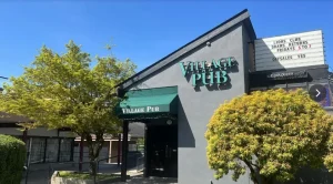 The exterior storefront of The Village Pub under a clear blue sky, featuring a green awning and signage for Lions Club draws and off-sales.