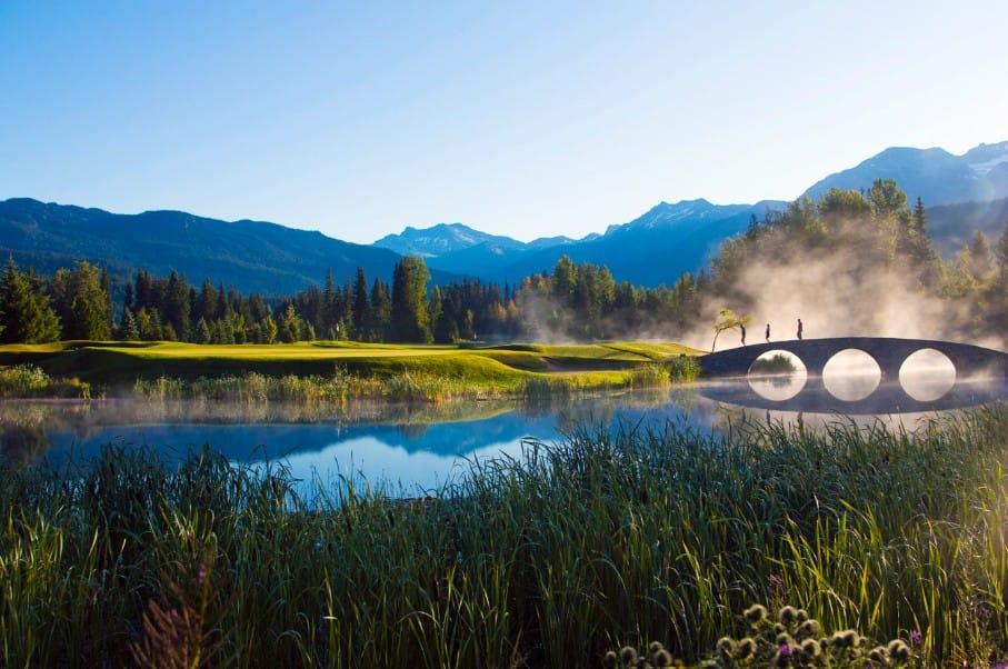 Two golfers crossing the stone bridge at Nicklaus North golf Whistler with morning mist rising off the lake and mountains in the background